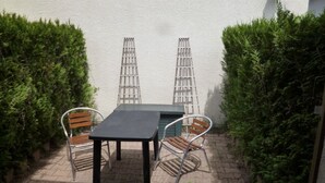 A patio with a table and two chairs, surrounded by neatly trimmed hedges and a white wall. - Seasonal rental (Les Mathes)