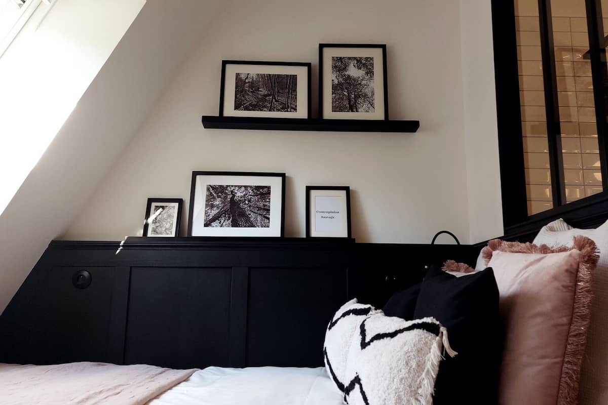 Un dormitorio con techo inclinado, tragaluz, cama con ropa de cama blanca, almohadas negras y blancas, y tres cuadros en un estante negro sobre la cama.