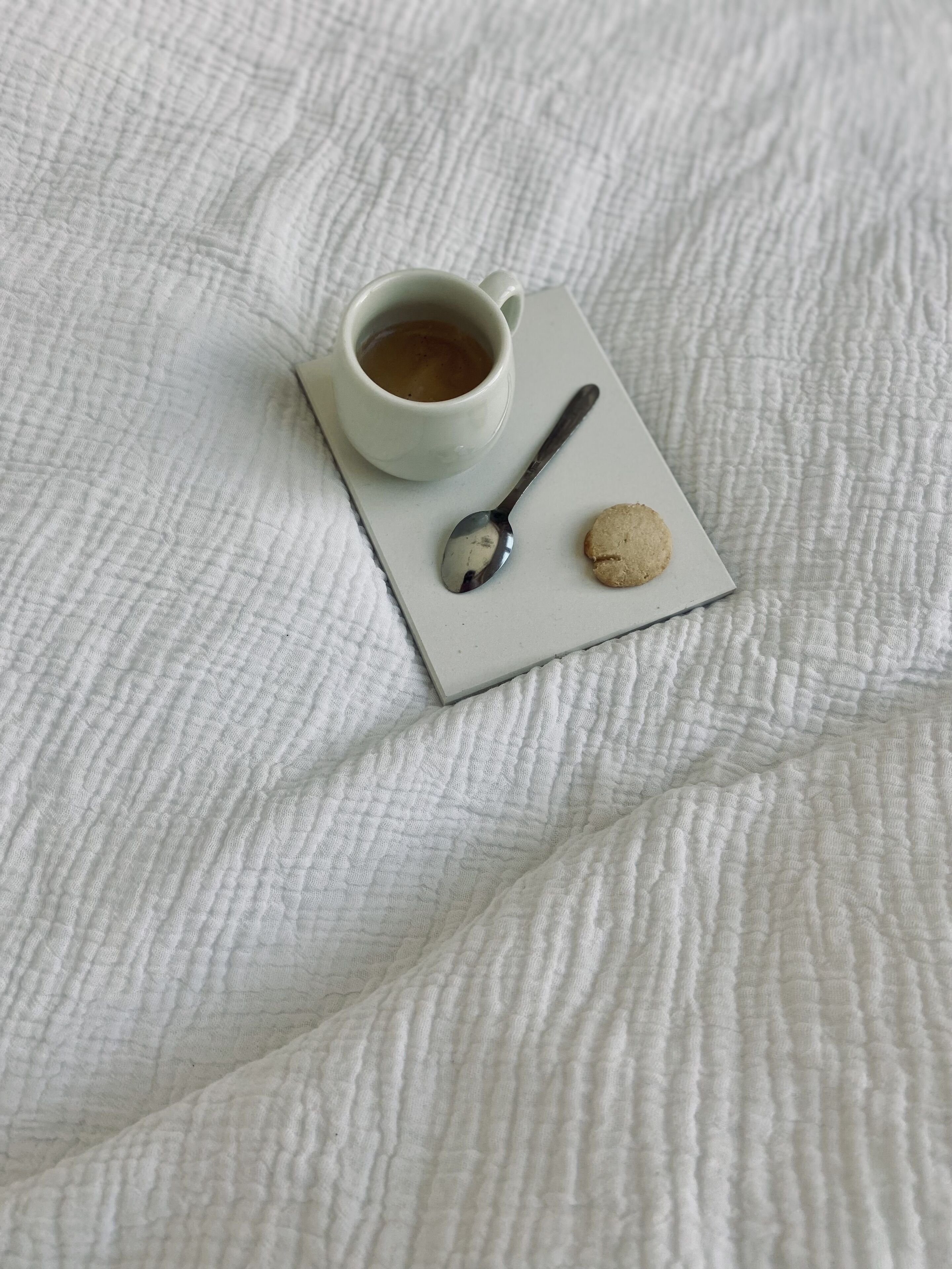 Una colcha blanca con textura y una bandeja blanca que sostiene una taza de café, una cuchara y una galleta.