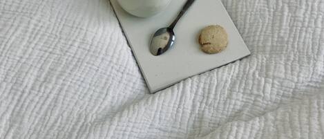 Una colcha blanca con textura y una bandeja blanca que sostiene una taza de café, una cuchara y una galleta.