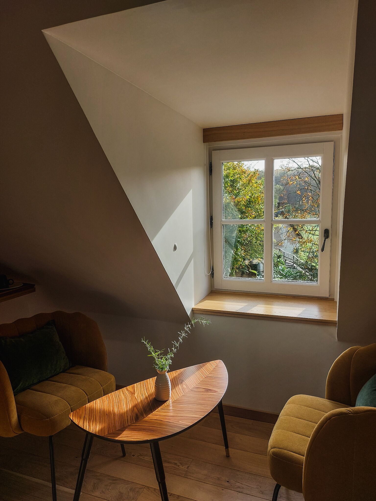 A room with a sloped ceiling, a window with a view of trees, a wooden table with a plant, and two chairs.