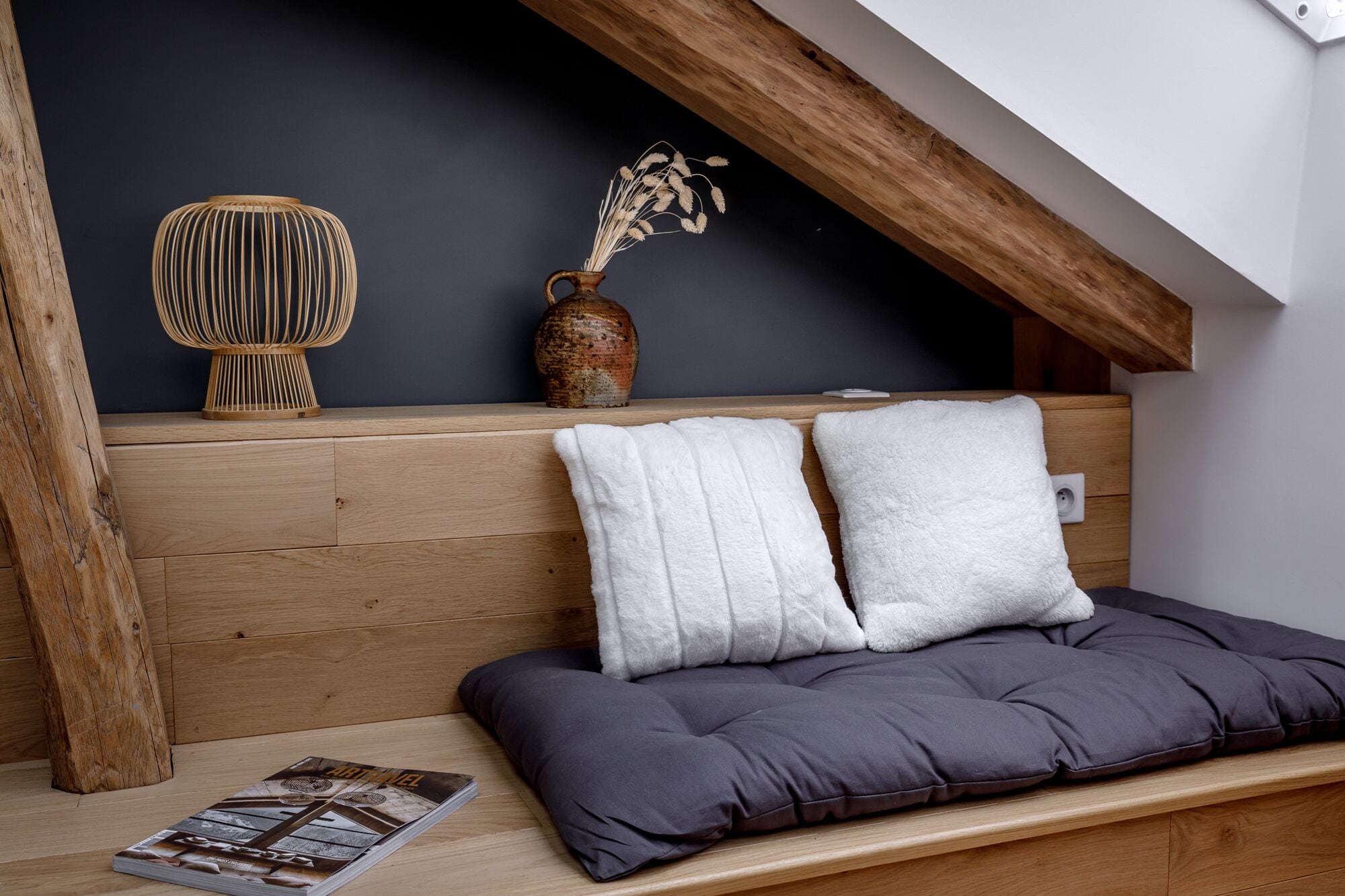 Une chambre à coucher confortable, aménagée en mezzanine, avec un plafond en pente en bois, une tête de lit en bois, un canapé gris avec des coussins, un panier en osier et un vase en céramique contenant des plantes séchées.