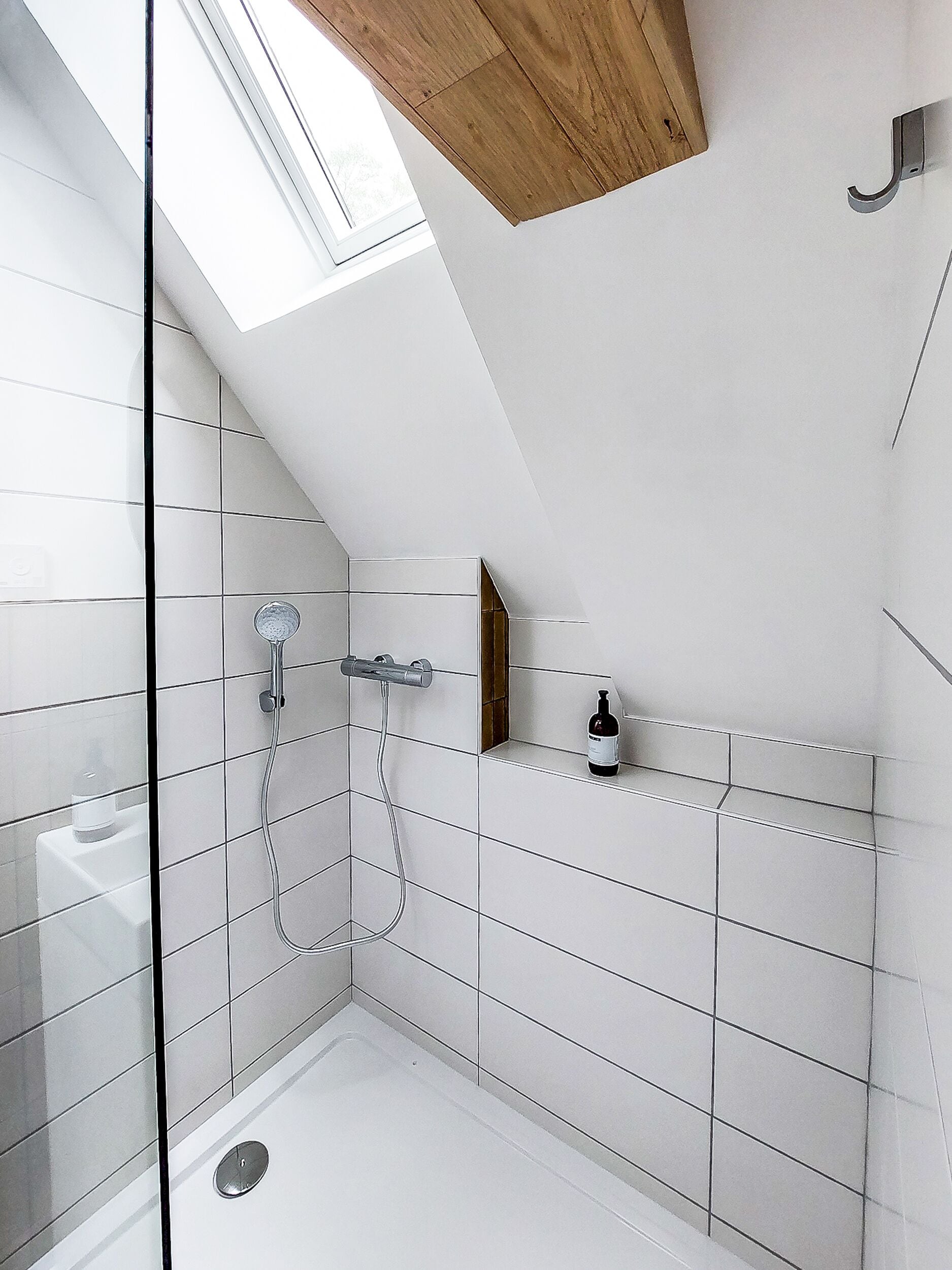 A modern shower with a glass door, white tiled walls, and a wooden ceiling beam.