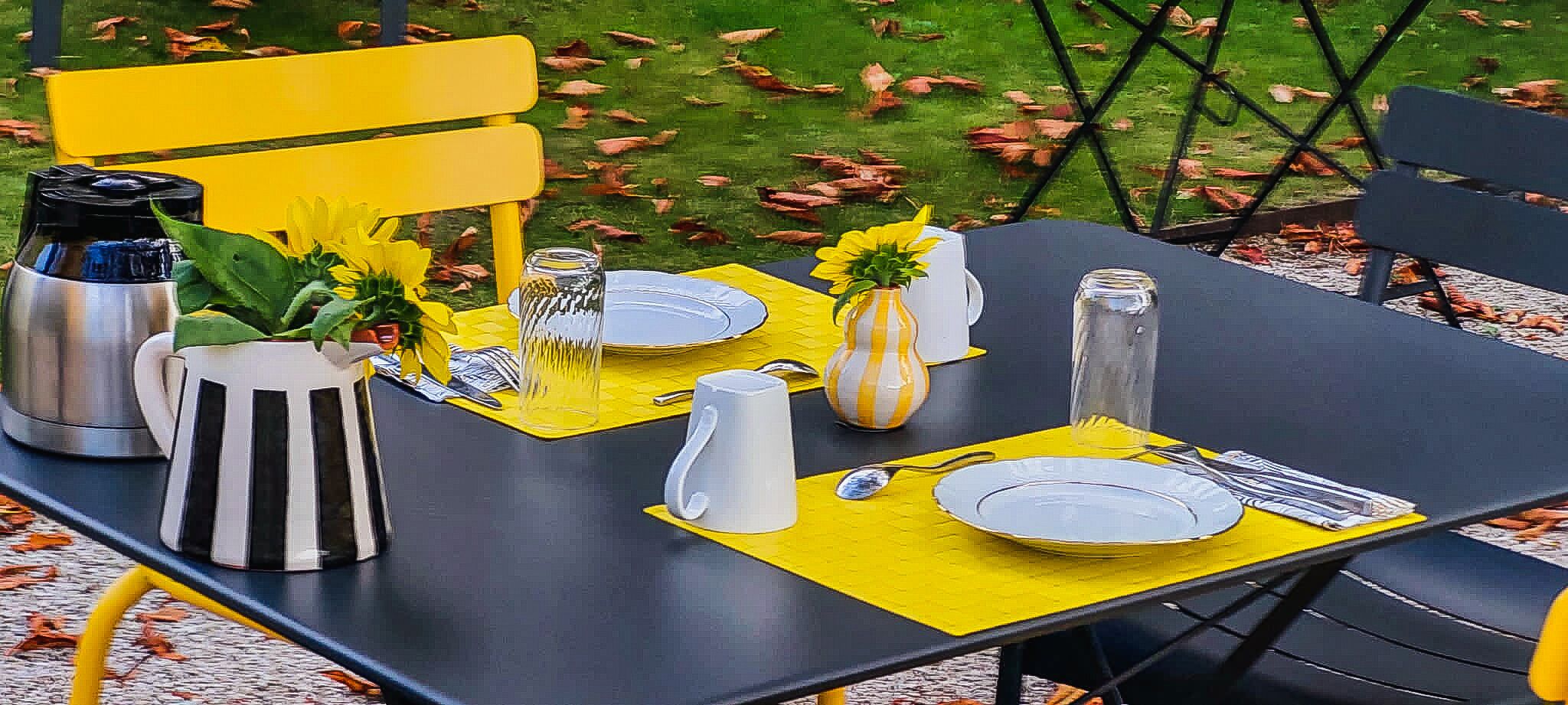 Une table dressée à l’extérieur, avec des sets de table jaunes, des assiettes blanches et divers types de verres.