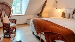 A cozy bedroom with a bed, a wicker chair, a bedside table with a lamp, and a window with a view of greenery. - Domaine du Val Sauvage (Langeais)