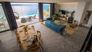 A spacious living area with a large window overlooking a pool and the sea. There is a dining table set with plates, glasses, and cutlery. A blue sofa and a TV stand with a flat-screen TV are also visible.