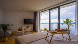 A modern bedroom with a large bed, a dining table with fruit, a TV, and a view of the sea.