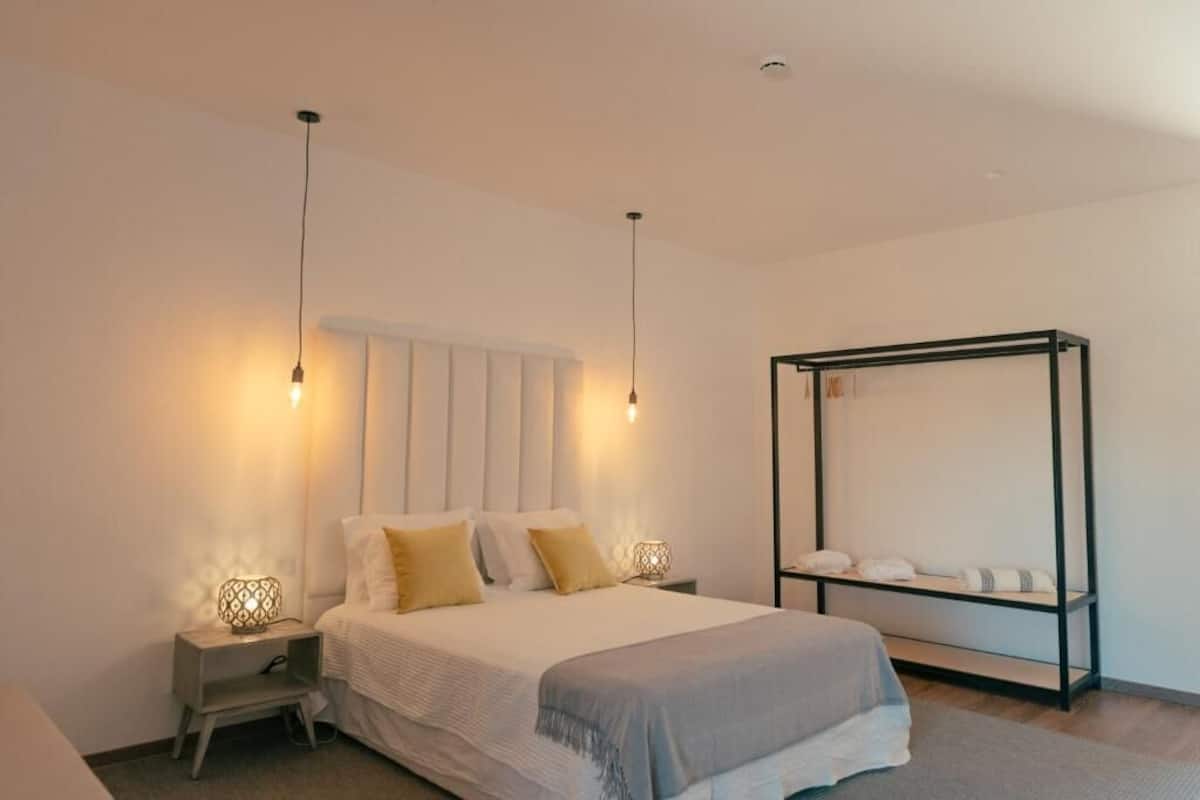 A bedroom with a bed, bedside lamps, a nightstand, a large mirror, and a shelf with towels.