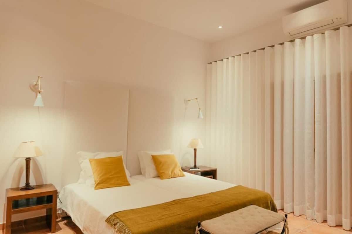A hotel room with a bed, two bedside tables with lamps, a window with vertical blinds, and a wall-mounted air conditioning unit.