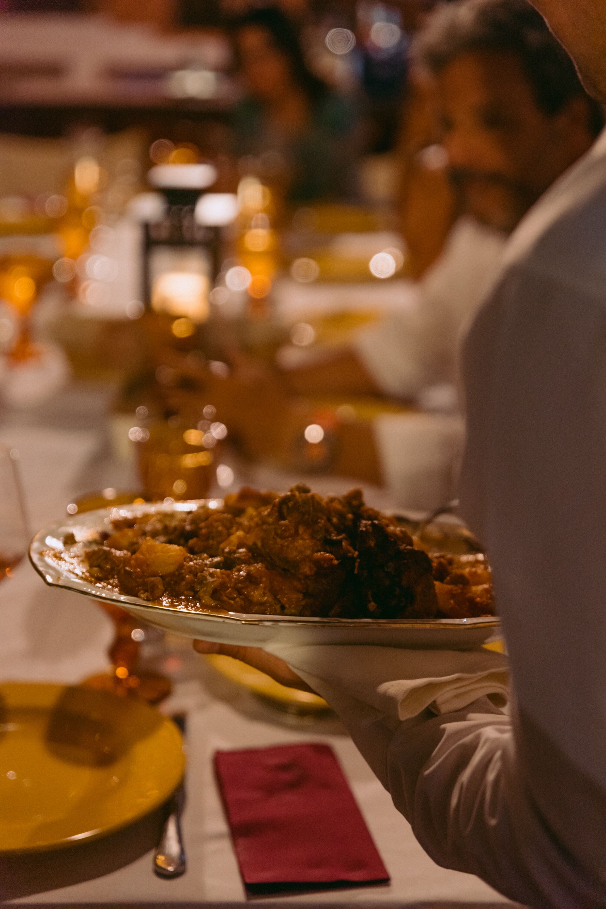A person serving a dish on a tray at a dining table with a red napkin and a spoon.