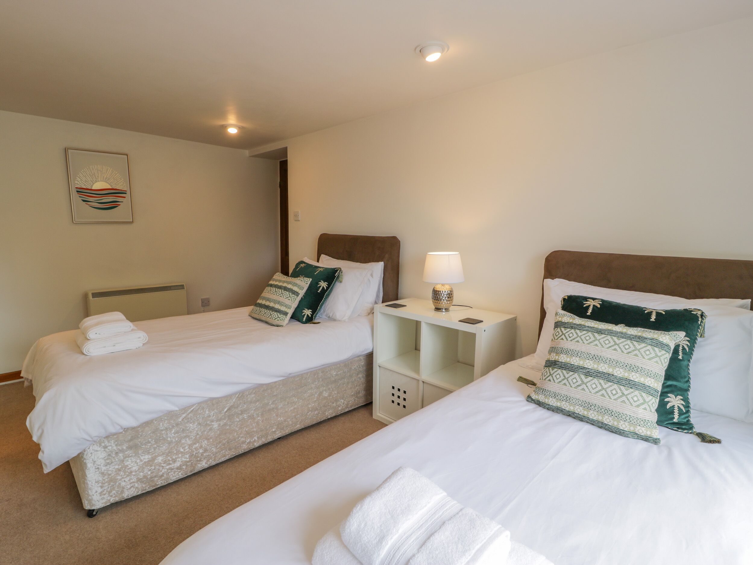 Habitación de hotel con dos camas, cada una con ropa blanca y almohadillas decorativas, una mesita de noche con lámpara, y un cuadro en la pared.