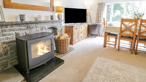 A cozy living room with a stone fireplace, a flat-screen TV, a dining area with wooden chairs, and a large window with curtains. - Bwthyn Pengwern (Caernarfon)