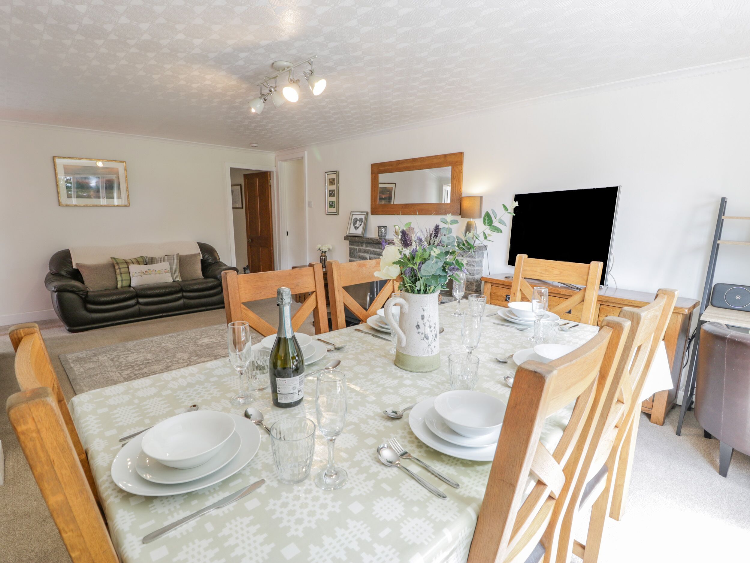 Un coin repas avec une table dressée pour six personnes, entourée de chaises en bois. On aperçoit un canapé et une cheminée au fond.