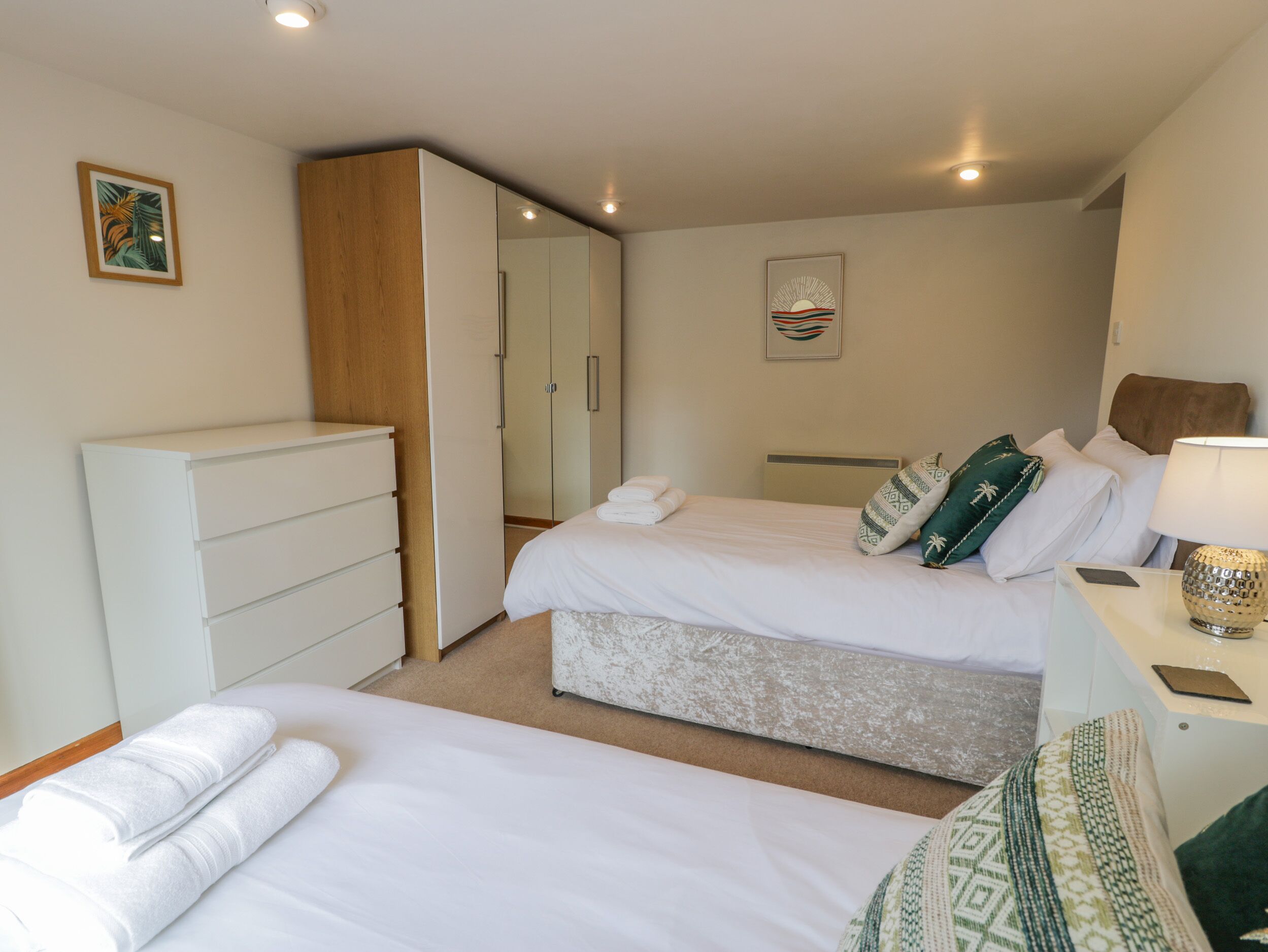 A hotel room with two beds, a wardrobe, a bedside table with a lamp, and a wall art piece.