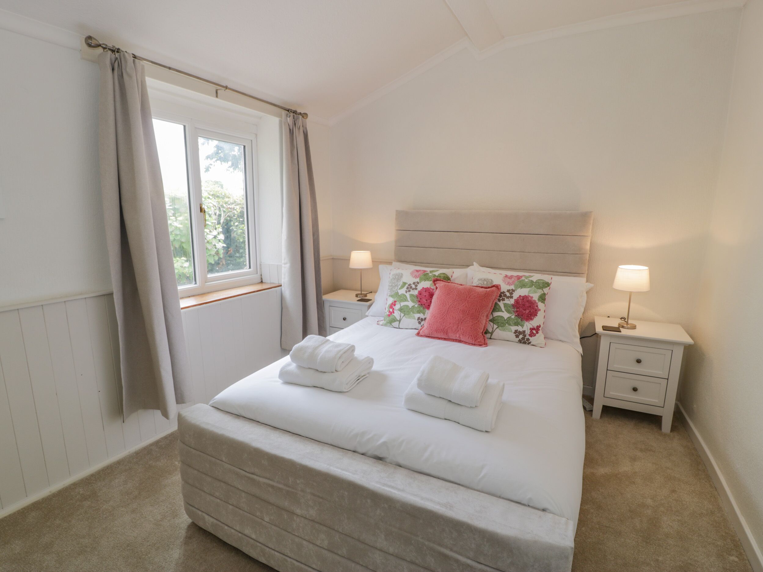 A neatly made bed with white linens and pink pillows, two bedside tables with lamps, a window with curtains, and a radiator under the window.