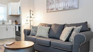 A modern living room with a grey sofa, patterned cushions, a round wooden coffee table, and a painting of trees on the wall. - Skip the hotel. Stay 4 min from Maxwell, walk to downtown & Civil Rights landmarks, and bring your dog. (Montgomery)
