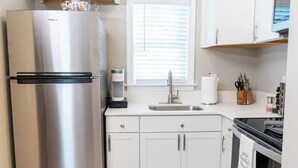 Une cuisine moderne dotée d’un réfrigérateur en acier inoxydable, de meubles blancs, d’un évier et d’une fenêtre avec des stores.