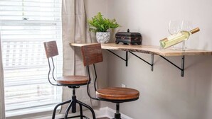 Un coin de pièce avec une fenêtre, deux tabourets de bar, une étagère avec une bouteille et des verres, et une plante en pot.