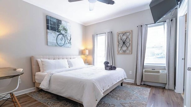 Une chambre à coucher comprenant un lit, un bureau, une chaise, un ventilateur de plafond, une télévision et une fenêtre avec des rideaux.