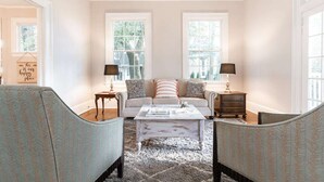 A living room with a sofa, two armchairs, a coffee table, and two lamps. There are two windows with curtains and a sign on the wall. - Made for the trips that bring everyone together. This historic Montgomery home gives families, wedding parties, and groups room to gather, room to disappear for a while, and one place to come back to—all minutes from downtown, Maxwell AFB, and the re (Montgomery)