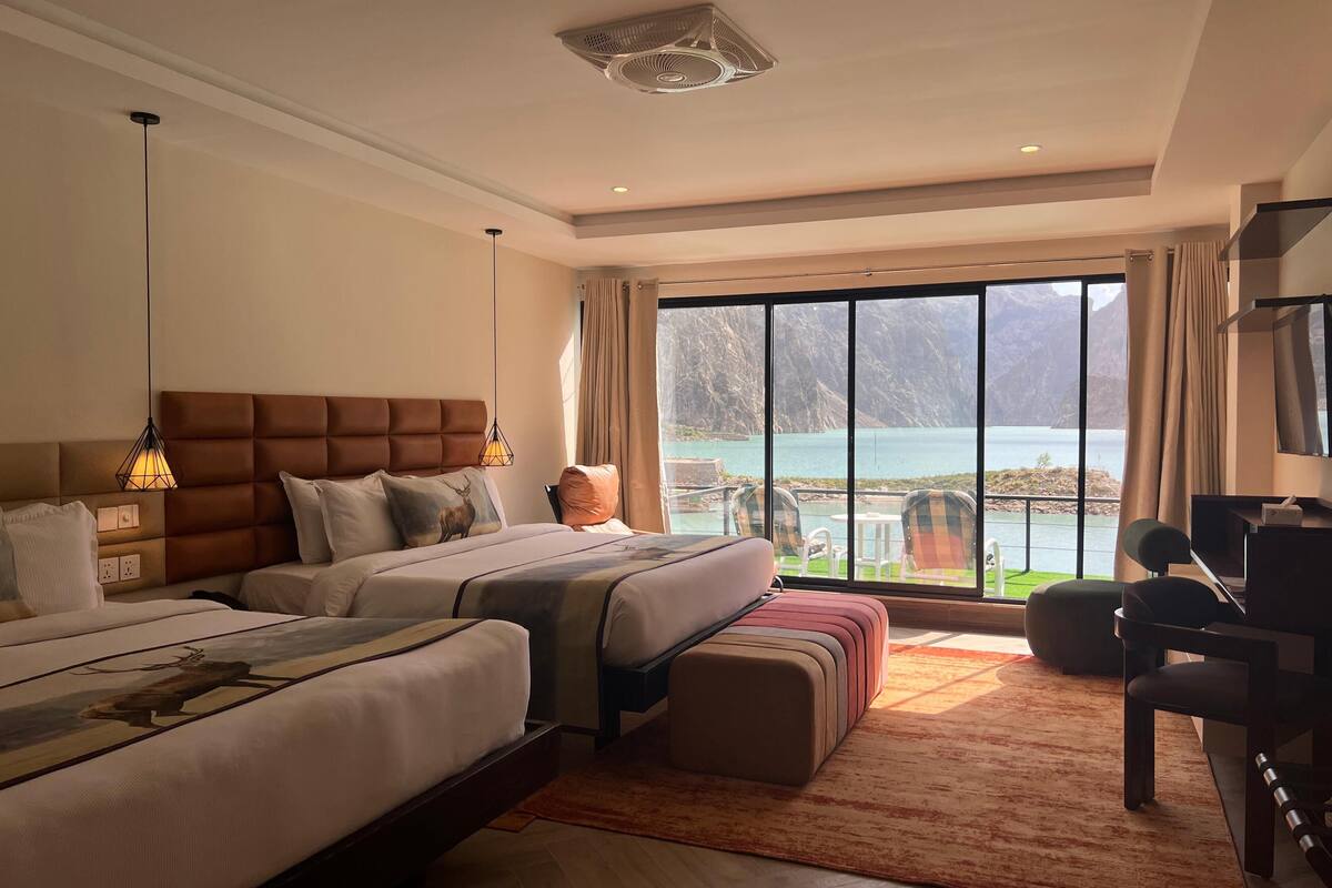 Habitación de hotel con dos camas, un ventanal con vistas a la montaña, un escritorio con televisor y un balcón con mobiliario exterior.