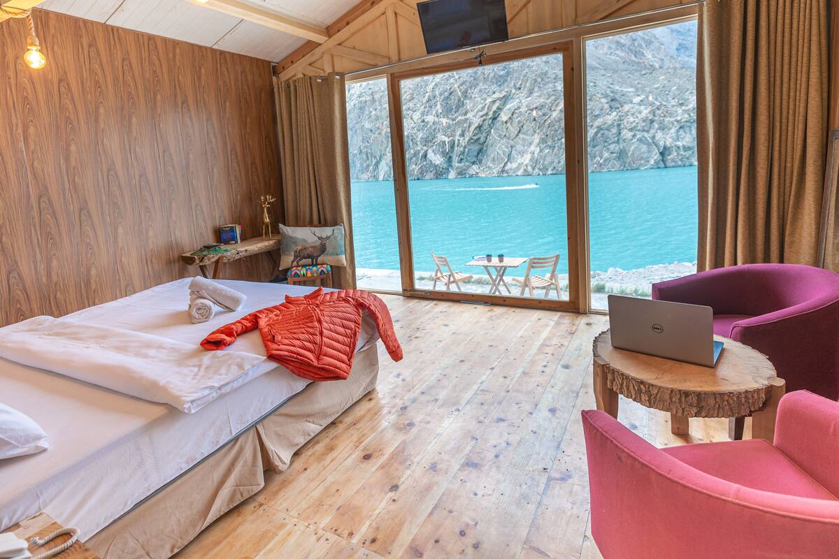 Una habitación con una cama, una laptop sobre una mesa de madera, una silla y vistas a un lago a través de amplios ventanales.