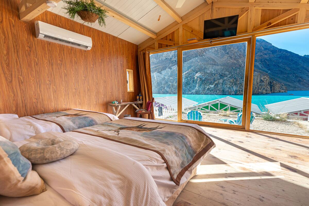 Dormitorio de cabaña de madera con una cama grande, aire acondicionado, televisión y vistas a aguas turquesas y montañas.