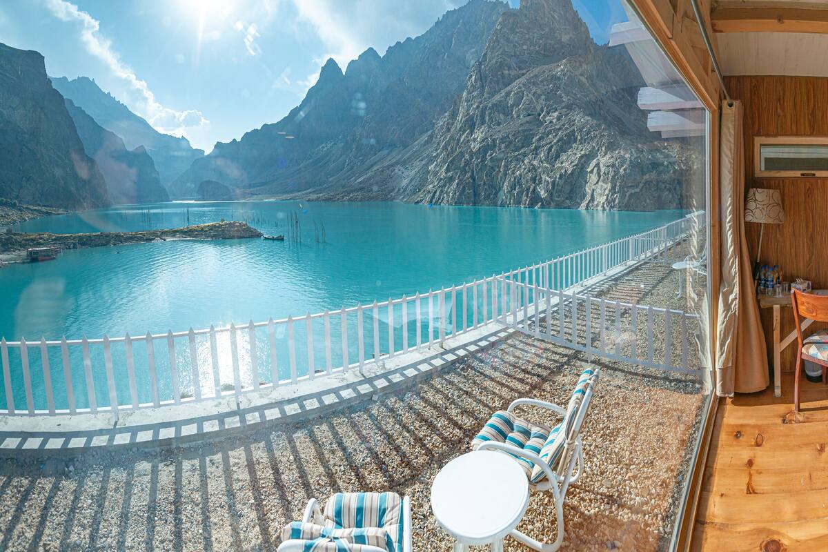 Terraza de madera con vistas a un lago turquesa y montañas, amueblada con sillas a rayas, una mesa redonda y una cama con ropa de cama estampada.