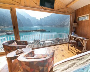 Une cabane en bois dotée d’un balcon donnant sur un lac et des montagnes. On y trouve deux chaises et une petite table avec des bouteilles.