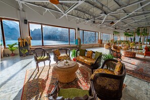 Une pièce spacieuse avec de grandes fenêtres donnant sur un lac et des montagnes. Le mobilier comprend des fauteuils et des canapés en osier, agrémentés de coussins.