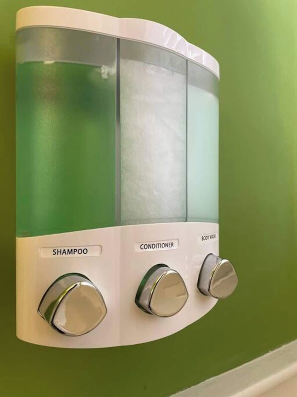 Eine wandmontierte Spenderstation mit drei Behältern, beschriftet mit Shampoo, Conditioner und Duschgel.