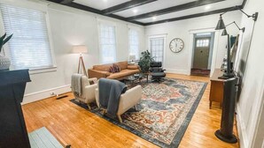 Un salon spacieux avec un grand tapis, un canapé, deux fauteuils et une table basse. Il y a une horloge au mur et une lampe sur une table d’appoint.