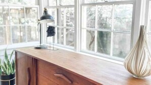 Une commode en bois avec des tiroirs et une lampe, placée près de grandes fenêtres donnant sur des arbres.