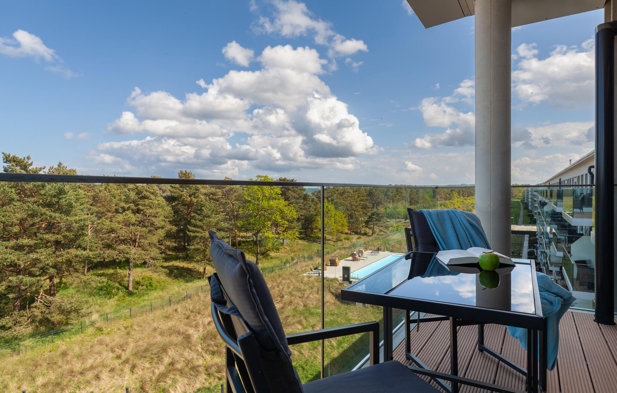 A balcony with a glass railing, a table with a chair, and a view of a forested landscape.