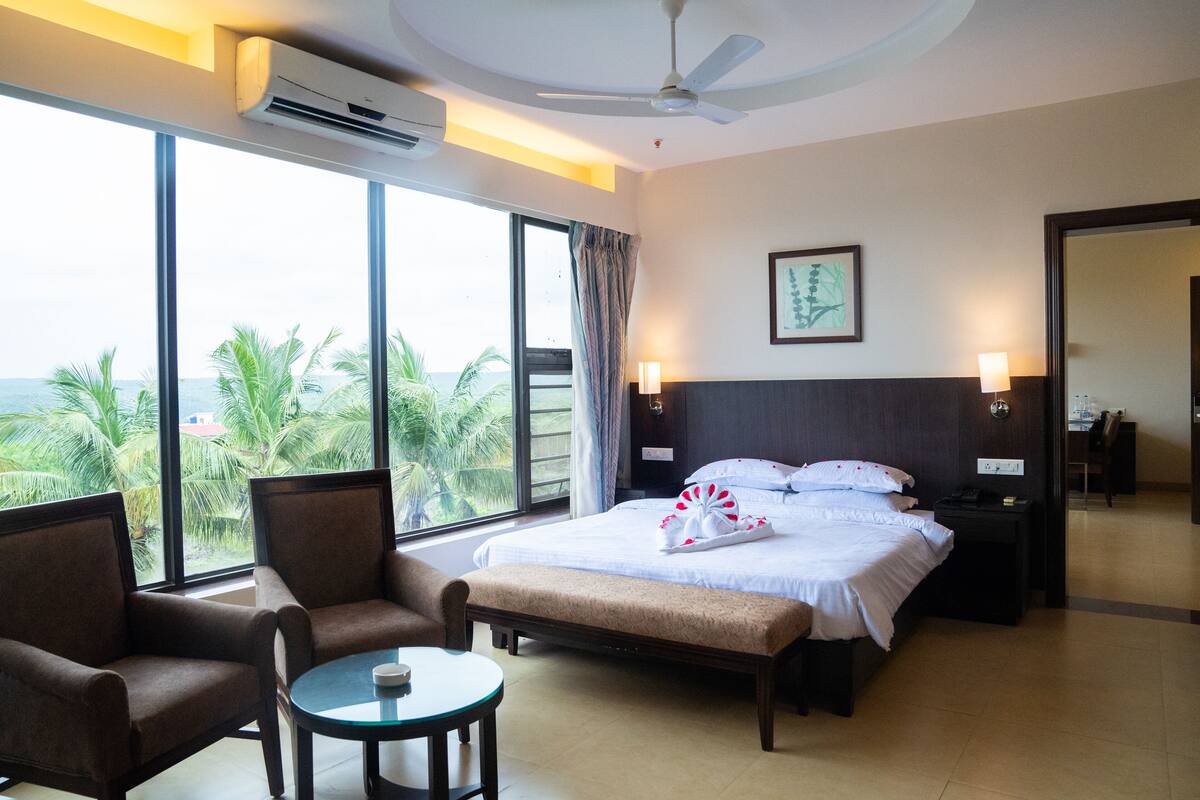 A hotel room with a bed, two armchairs, a small table, and a large window offering a view of palm trees and the sea.
