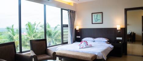 Habitación de hotel con una cama, dos sillones, una mesita y un ventanal grande que ofrece vistas a palmeras y al mar.