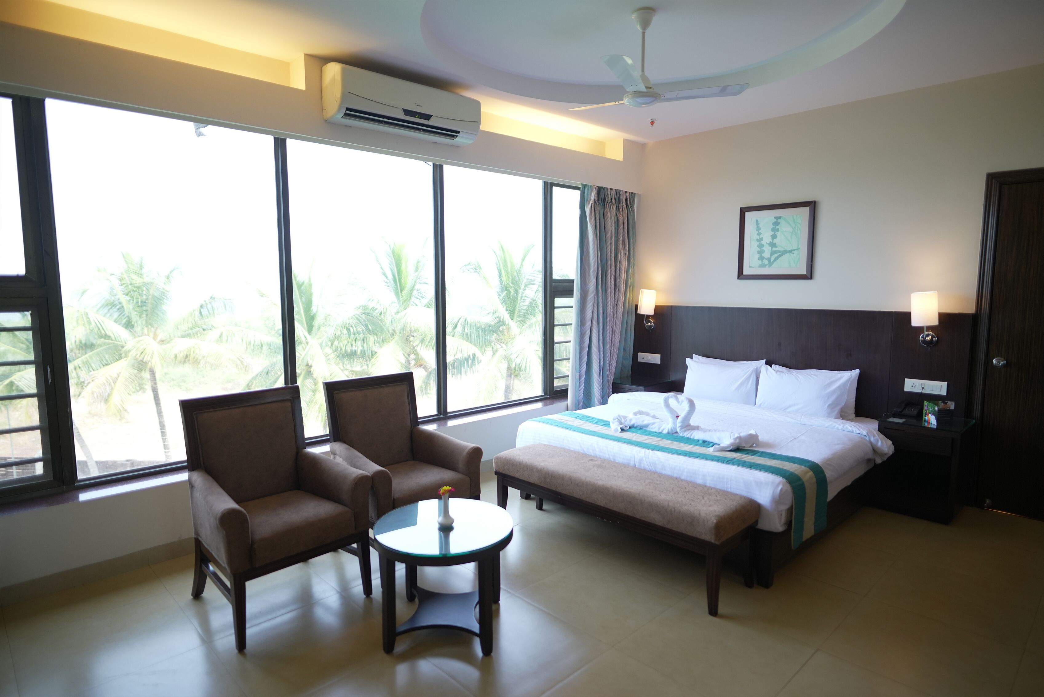 Habitación de hotel con una cama, dos sillones, una mesita y vistas a palmeras a través de amplias ventanas.