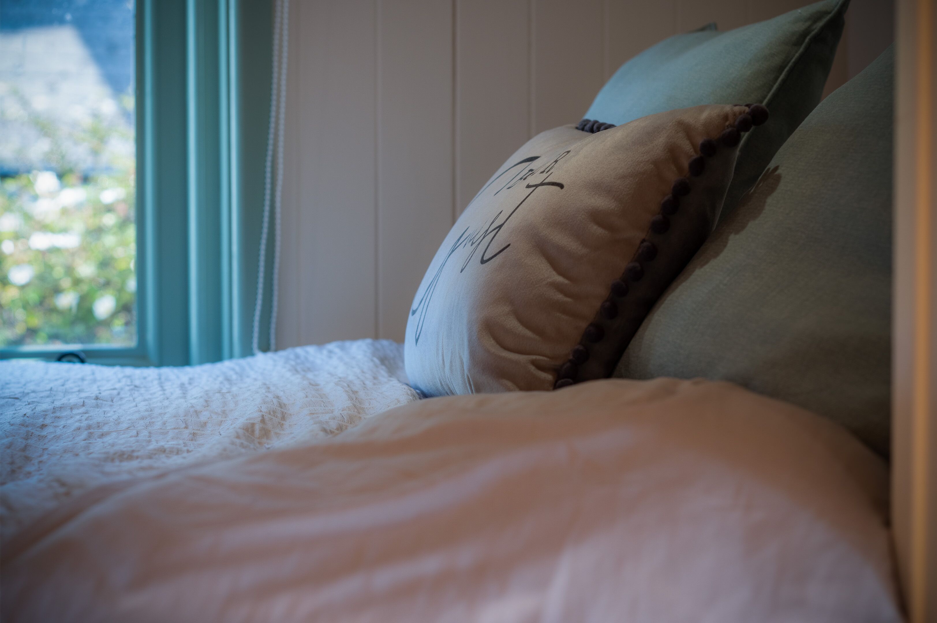 Una cama con una colcha blanca y dos almohadas, una con un diseño distintivo y la otra lisa, en una habitación con ventana.