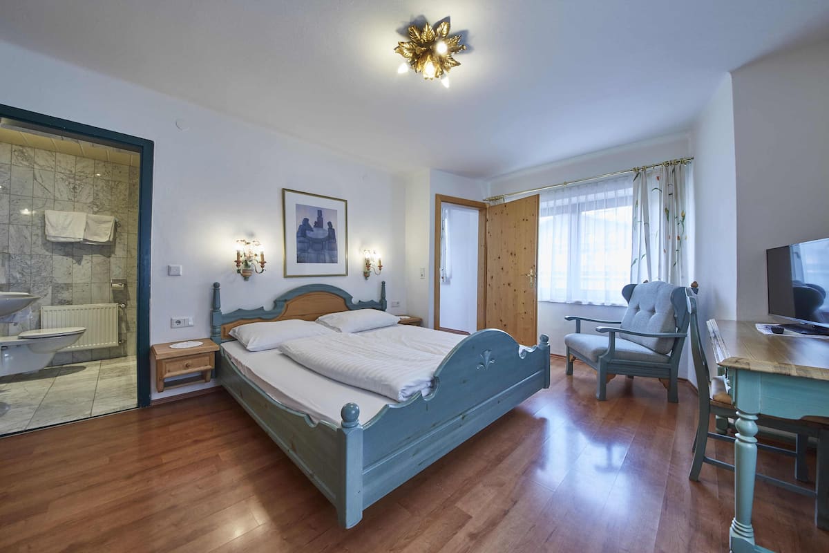 Une chambre à coucher comprenant un lit, une chaise, un bureau et une salle de bain visible à travers une porte vitrée.