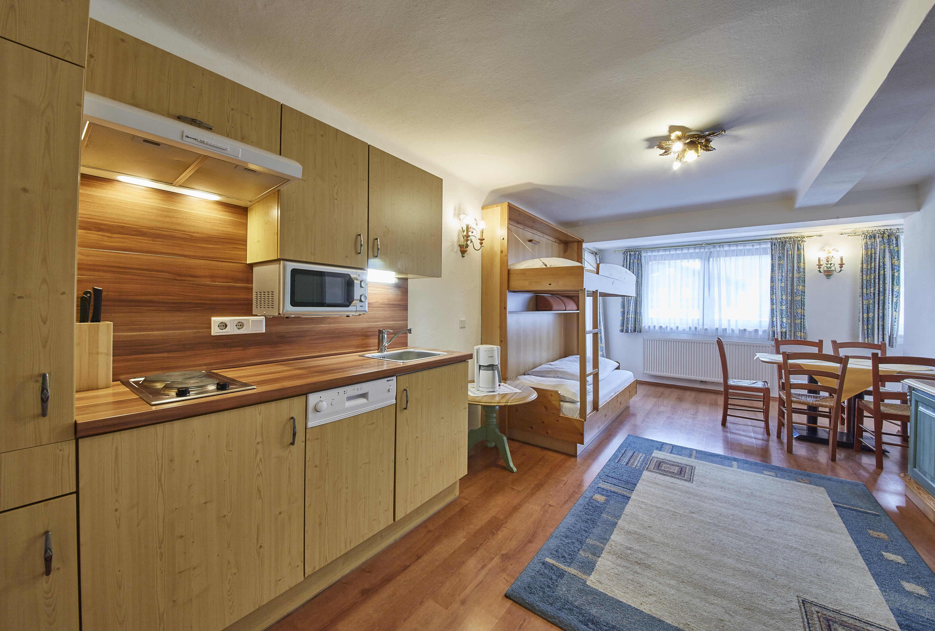 Cocina compacta con armarios de madera, un microondas y un lavabo. Hay un comedor con mesa y sillas, y una cama litera al fondo.
