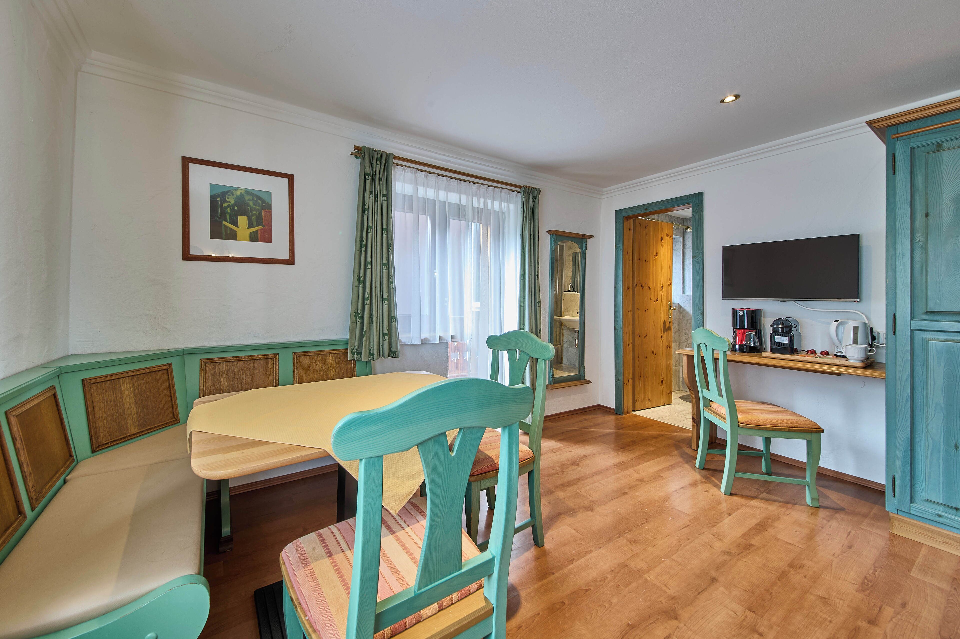 A dining area with a wooden table and chairs, a television mounted on the wall, and a window with curtains.