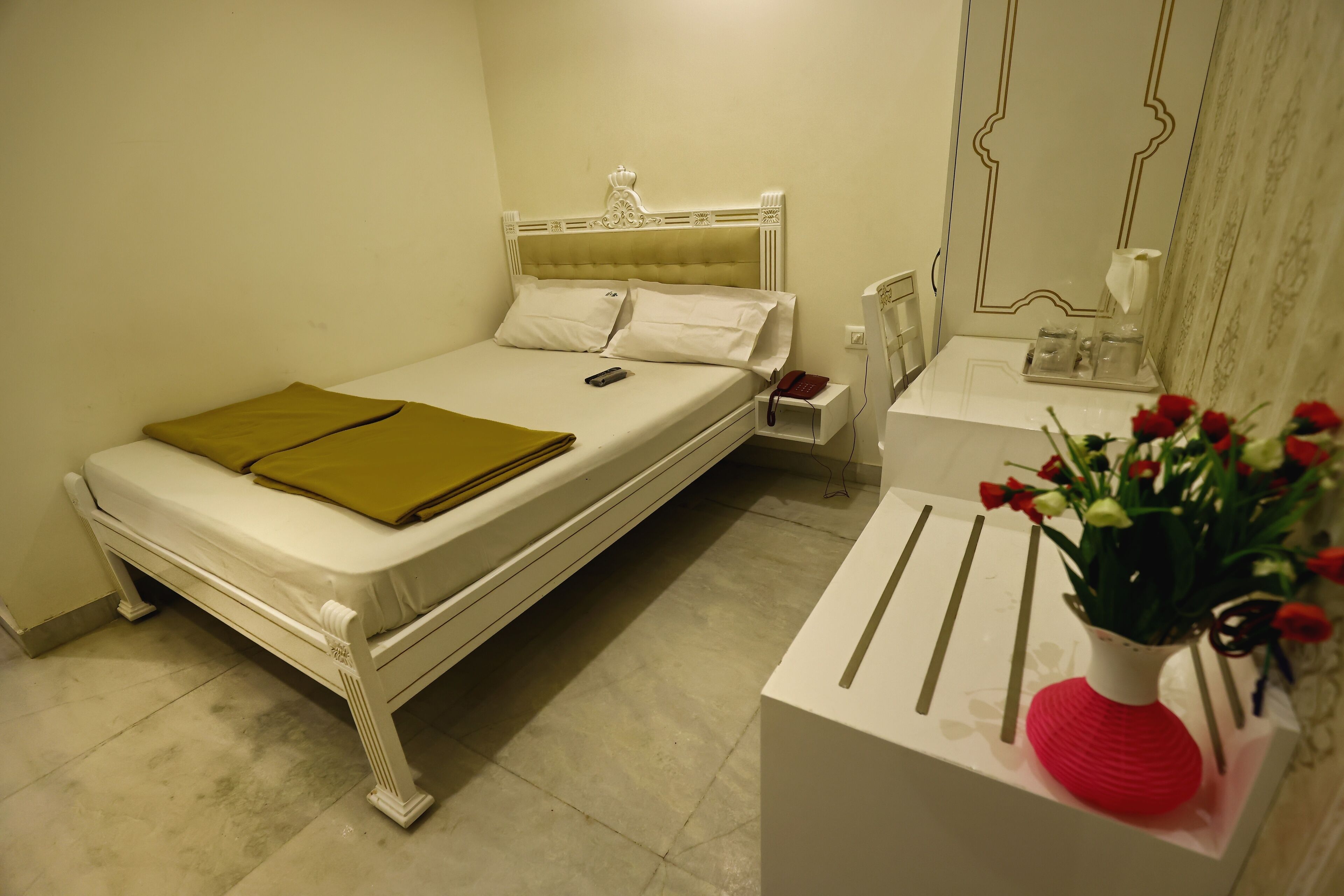 Una cama bien hecha con cabecera blanca, una mesita de noche con un florero y un pequeño tocador con un teléfono.