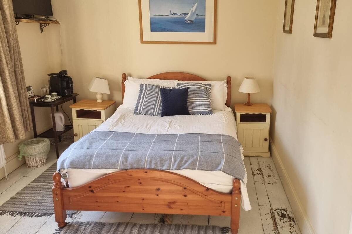 A wooden bed with a grey and white checkered blanket, two bedside tables with lamps, a framed picture on the wall, and a small table with a kettle and cups.