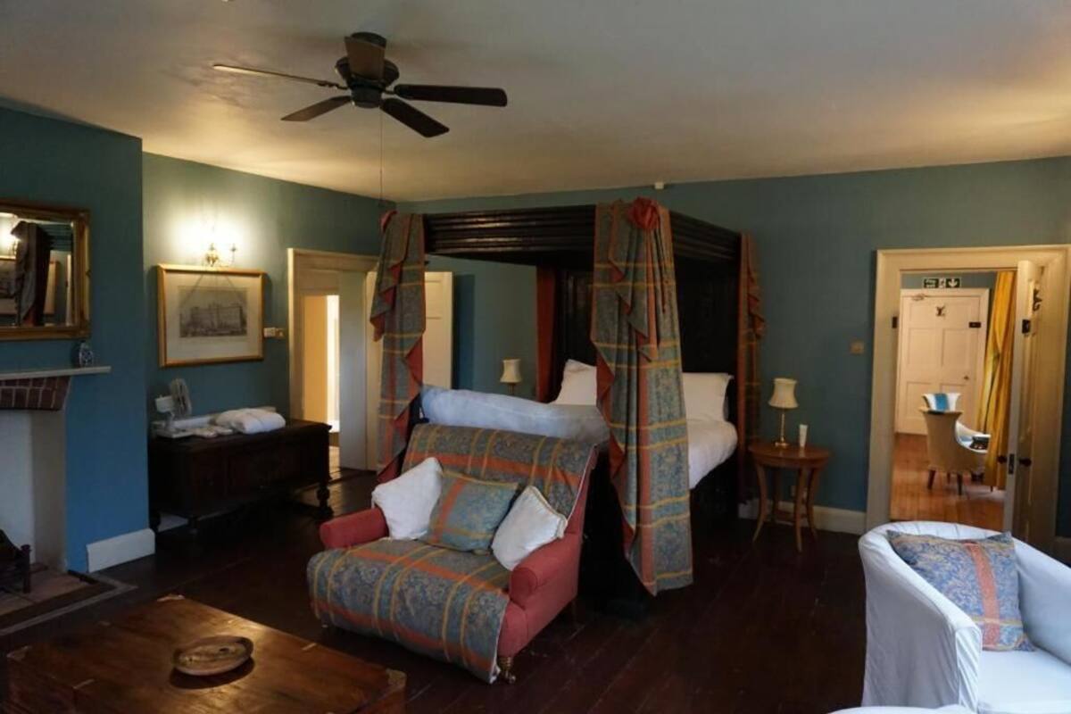 A room with a four-poster bed, a red armchair, a white armchair, a small table, and a ceiling fan.