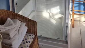 Une douche fermée par des parois de verre, avec un mur en briques blanches, une chaise en bois et un panier en osier contenant des serviettes.