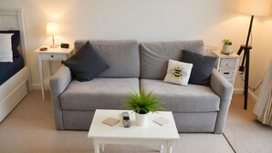 A neutral-colored living room with a grey sofa, a small white table with a plant, a remote control, and decorative pillows. - Lodge 7 - Lytton Tree Lodge, Reydon, Southwold (Reydon)