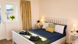 A neatly made bed with a blue bedspread, two pillows, and a welcome pillow. There are two bedside tables with lamps and a small plant. - Lodge 7 - Lytton Tree Lodge, Reydon, Southwold (Reydon)