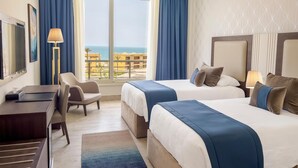Ein Hotelzimmer mit zwei Betten, einem Schreibtisch, einem Sessel und Meerblick.