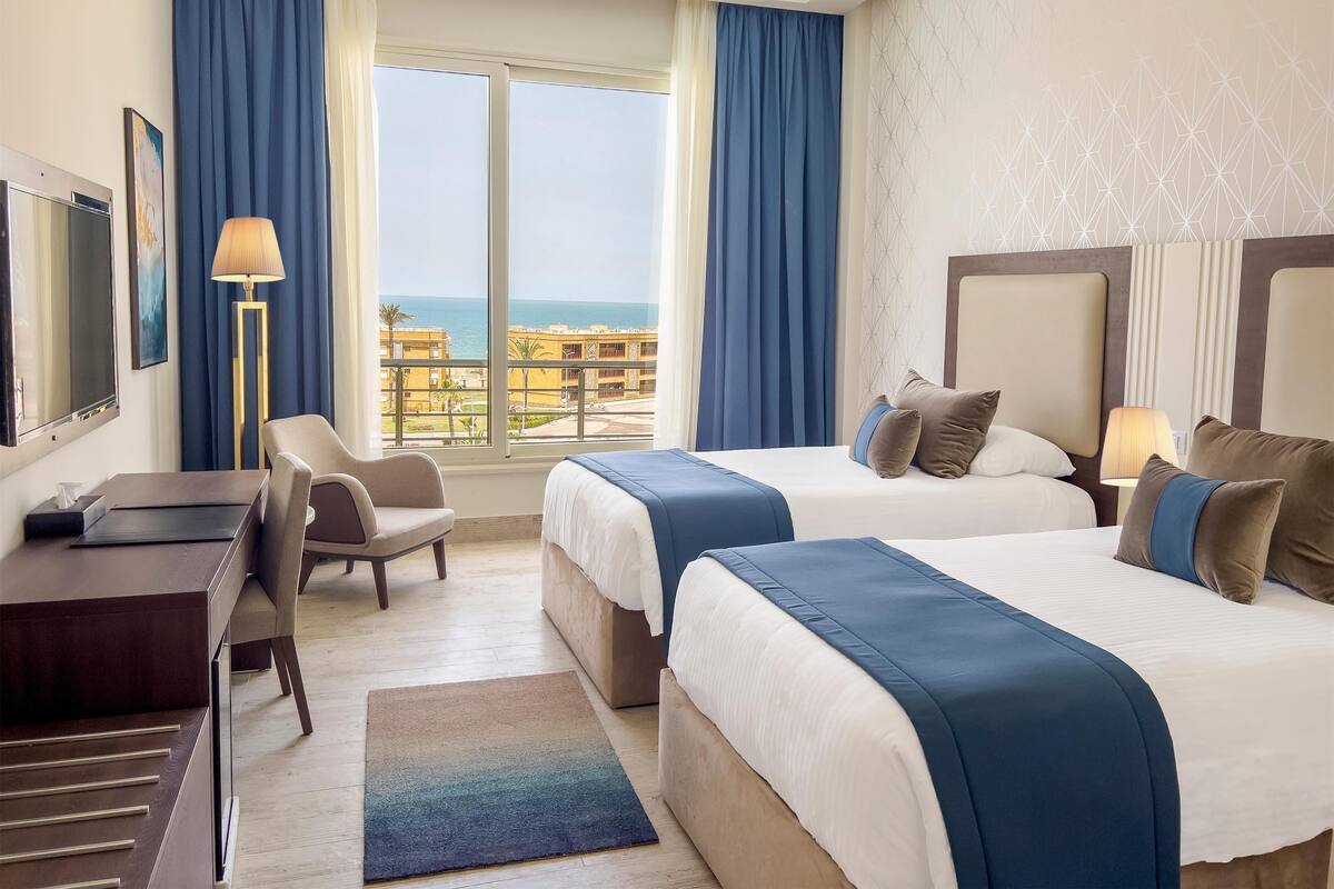 A hotel room with two beds, a desk, a chair, a lamp, and a view of the sea.