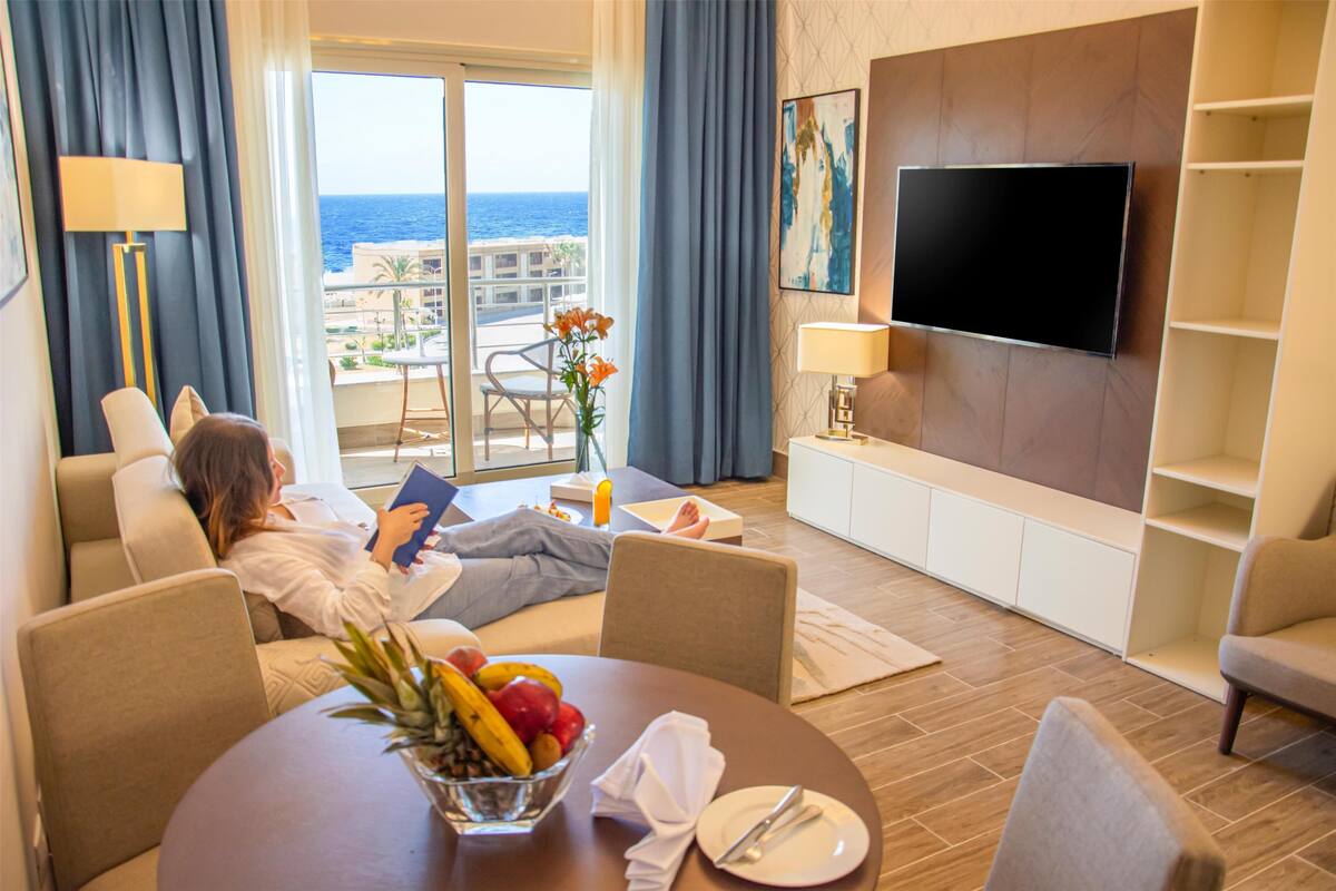 A hotel room with a balcony, a flat-screen TV, a dining area with a fruit bowl, and a view of the sea.